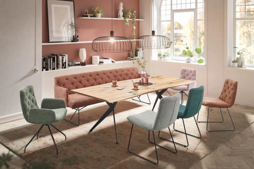 Esszimmer romantisch seca Esszimmerstuehle in Cord , verschiedene Farben mintgruen, gruen, orange, rosa. Holztisch in der Mitte . Hinter Essgruppe eine Terrakotawand mit eingebauten Holzregalen.