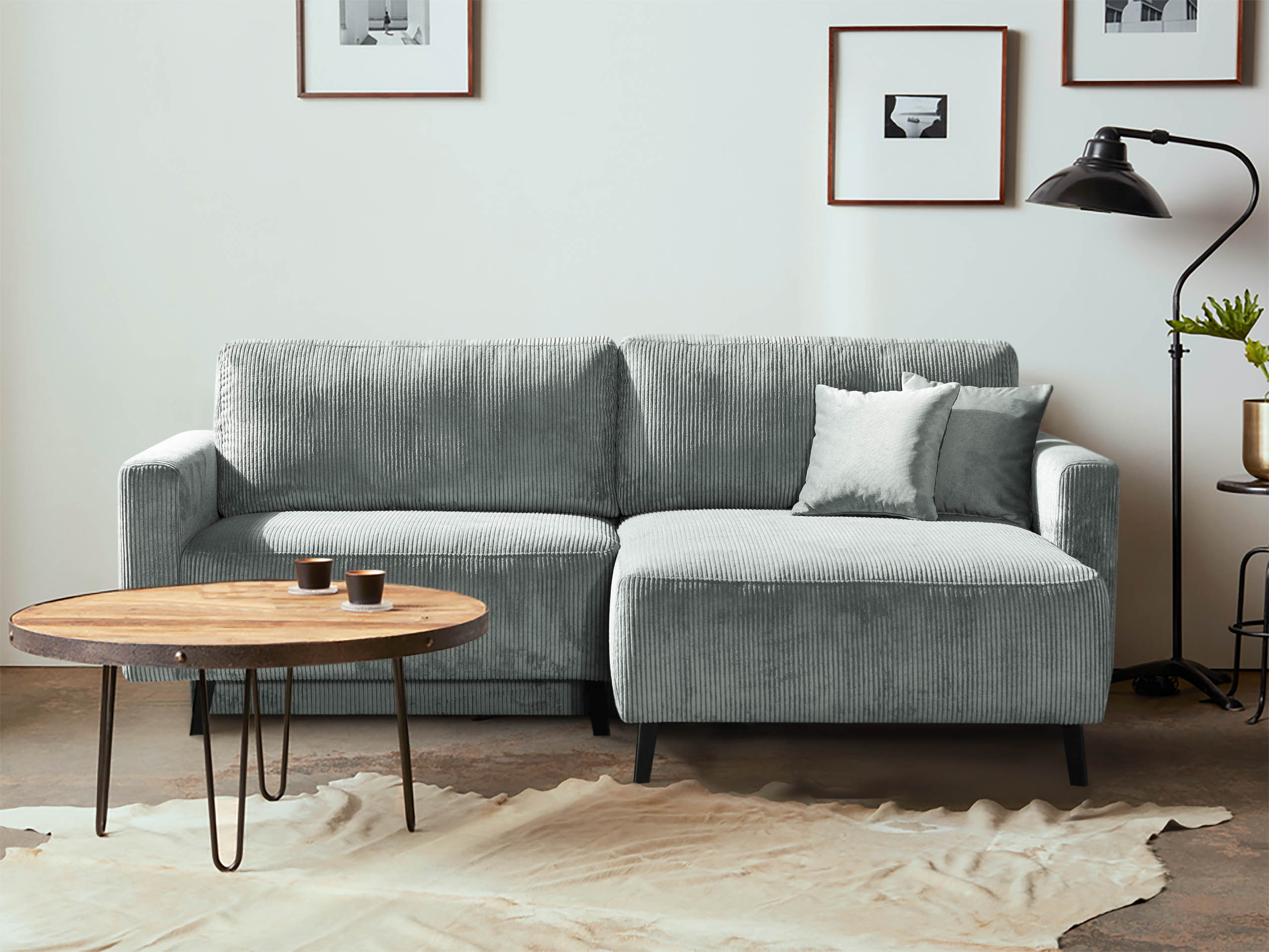ein kleines ecksofa - schlafsofa in cord grau steht an der wand. gewebter naturfarben teppich liegt davor. ein runder couchtisch steht vor dem Sofa mit metallfüßen und holzplatte.