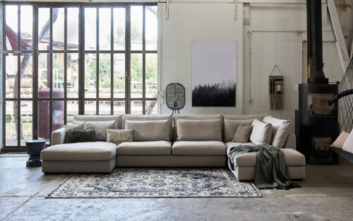 loft werkgebäude eine beige wohnlandschaft steht in der mitte. Dahinter riesen fenster und rechts ein metall kamin. teppich in der mitte