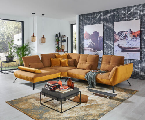 eine ecksofa mustard in geschwungenenen üppigen formen auf schwarzen hohen metallfüßen. an der seite schwarze wand mit zwei riesenbilder. vor dem sofa teppich in senf - grauen farben und ein schwarzer metall couchtisch