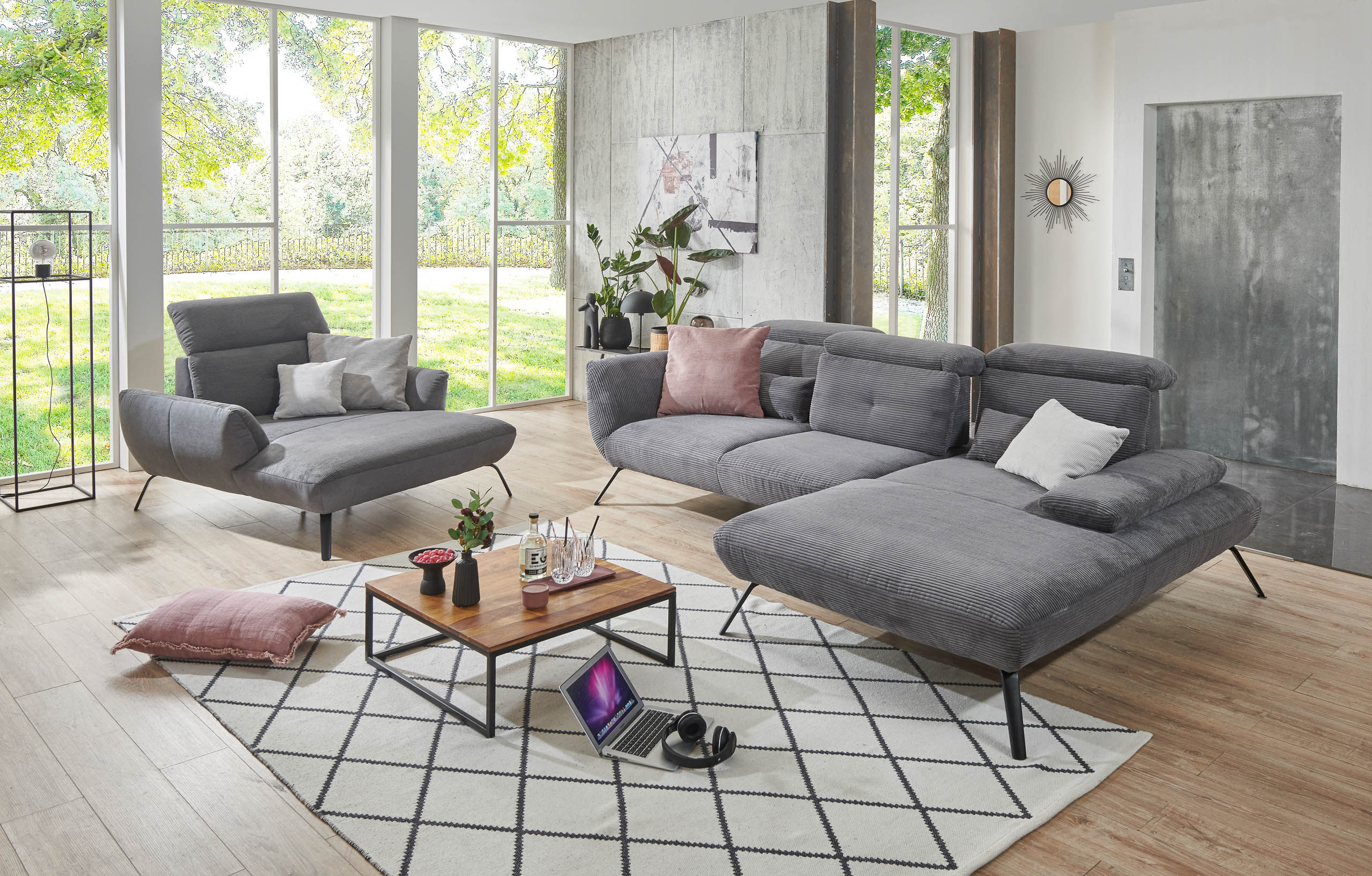 eine ecksofa in grau cord. sofa ist üppig hat geschwungene formen mit eingebauten kopfstützen verstellbar. steht auf chwarzen metallfüßen mit viel bodenfreiheit. steht i einem großen zimmer daneben ein loveseat in sofadesig. in der mitte ein couchtisch auf metallfüßen in schwarz und holzplatte. ein weißer teppich mit schwarzen rautenmuster liegt auf dem boden